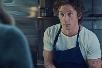 Screen capture of a character from the TV show 'The Bear' wearing a white t-shirt with a black apron over top.