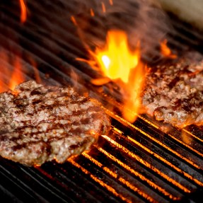 Two burgers on a grill, flames leaping up around them. They have char marks on them.