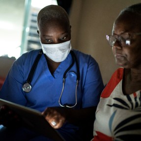 A health care provider wearing blue scrubs and a mask with a stethoscope around their neck looking at a screen with a senior hospital patient.