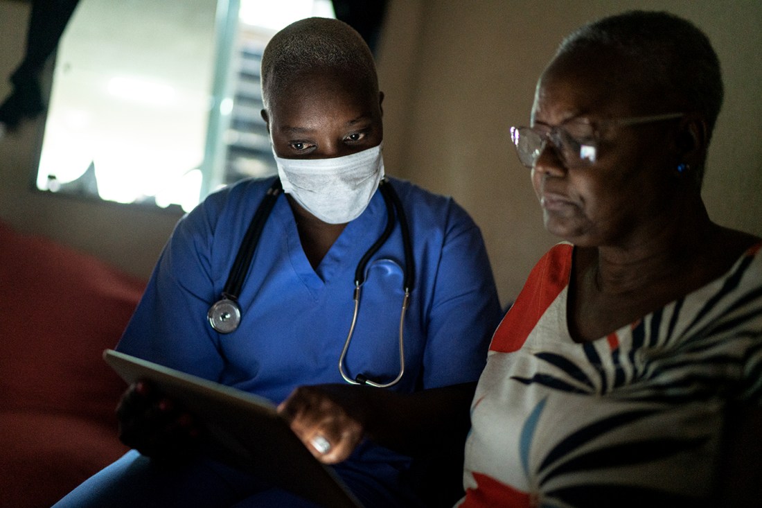 A health care provider wearing blue scrubs and a mask with a stethoscope around their neck looking at a screen with a senior hospital patient.