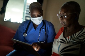 A health care provider wearing blue scrubs and a mask with a stethoscope around their neck looking at a screen with a senior hospital patient.