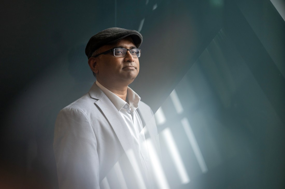Portrait of Sudhakaran Prabakaran wearing a white collared shirt underneath a grey blazer and a hat. 