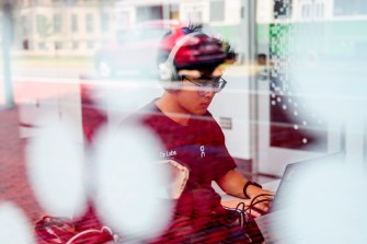 A person sits at a table using a laptop, seen through a window with reflections and soft light.