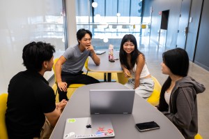 Four students sit around a table while looking at a laptop.