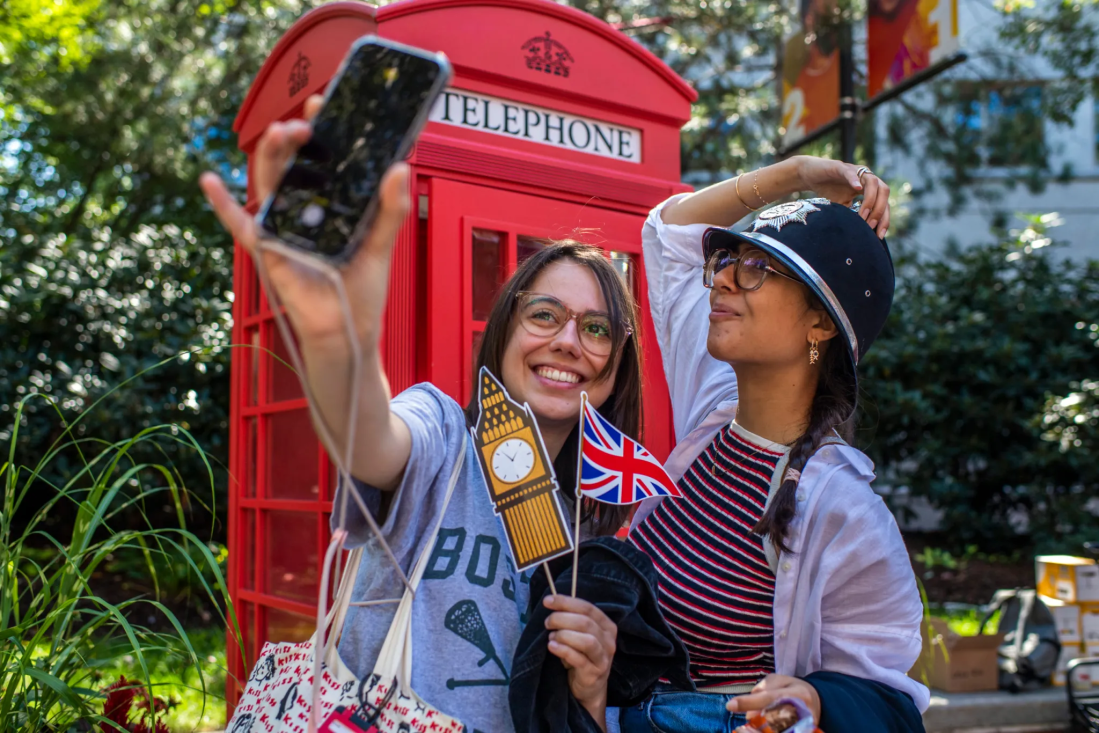 Two students posing for a selfie in front of a red telephone box holding a mini Union Jack and Big Ben cutouts in front of them