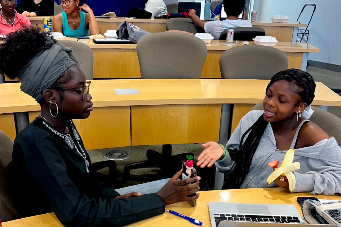 Two students sit across from each other. One is holding their phone up to the other and the other is gesturing to the phone with one hand and holding a banana in the other.