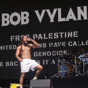 Bob Vylan wearing white shorts and no shirt singing into a microphone while lifting one leg in preparation to stomp on stage at Glastonbury.