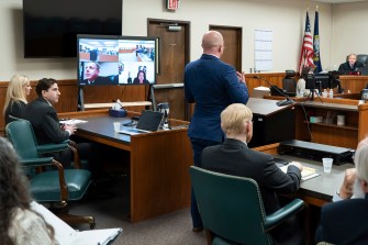 Jay Logsdon, center, an attorney representing Bryan Kohberger, second from left, standing in a court room speaking. In the background there is a screen with several people calling into the hearing.