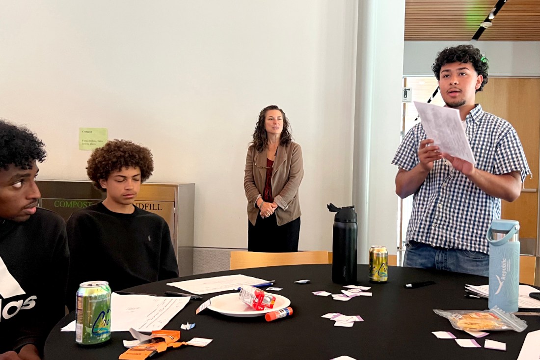 A student standing up next to a table holding a piece of paper in front of him and speaking to a room full of other students and at eacher.