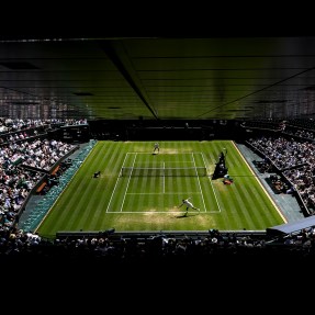 Aerial view of a tennis court during a match, showing the grass court from above with two players positioned on opposite sides. The court is surrounded by packed stadium seats filled with spectators.