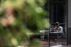 A student wearing headphones works on a laptop while seated at a table inside a glass-walled study space. The photo is taken from outside through a window, with blurred green foliage in the foreground.