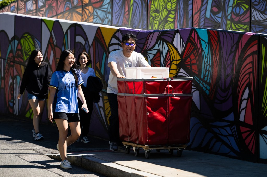 Three people walk along a sidewalk, while another person pushes a large hamper full of move-in belongings.
