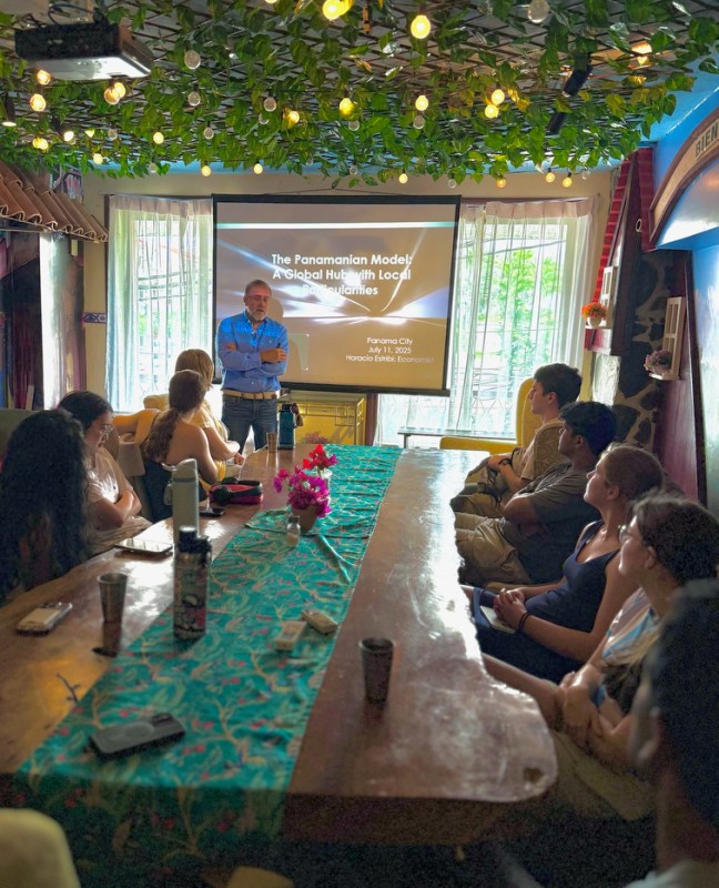 Students sitting at a large wooden table paying attention to a presenter in a blue shirt standing in front of a screen that says 'The Panamanian Model' as the title. 