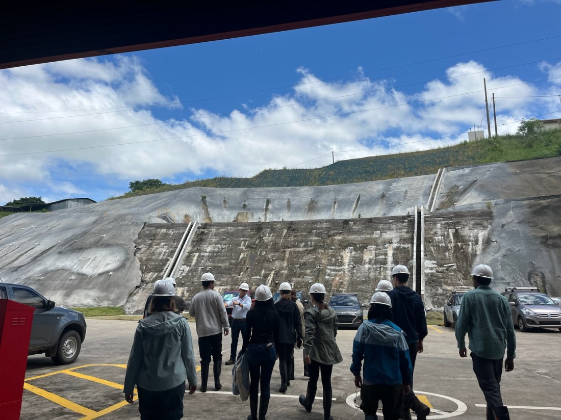 A group of people wearing white hard hats walks away from the camera across a paved area towards a large concrete dam structure.