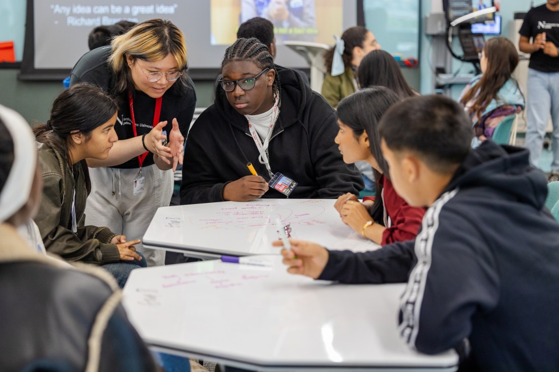 Students huddle together, brianstorming, over a white board with writing on it.