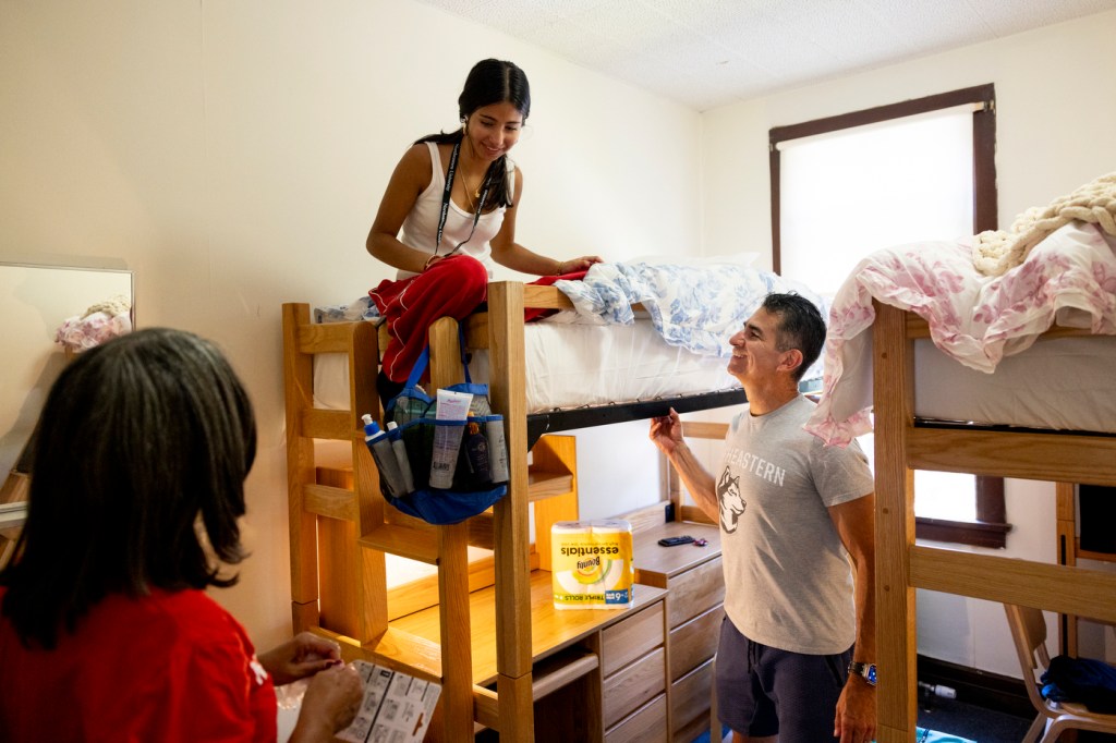 A student sits on top of a bunk bed unpacking while their family members stand in their dorm room with them.