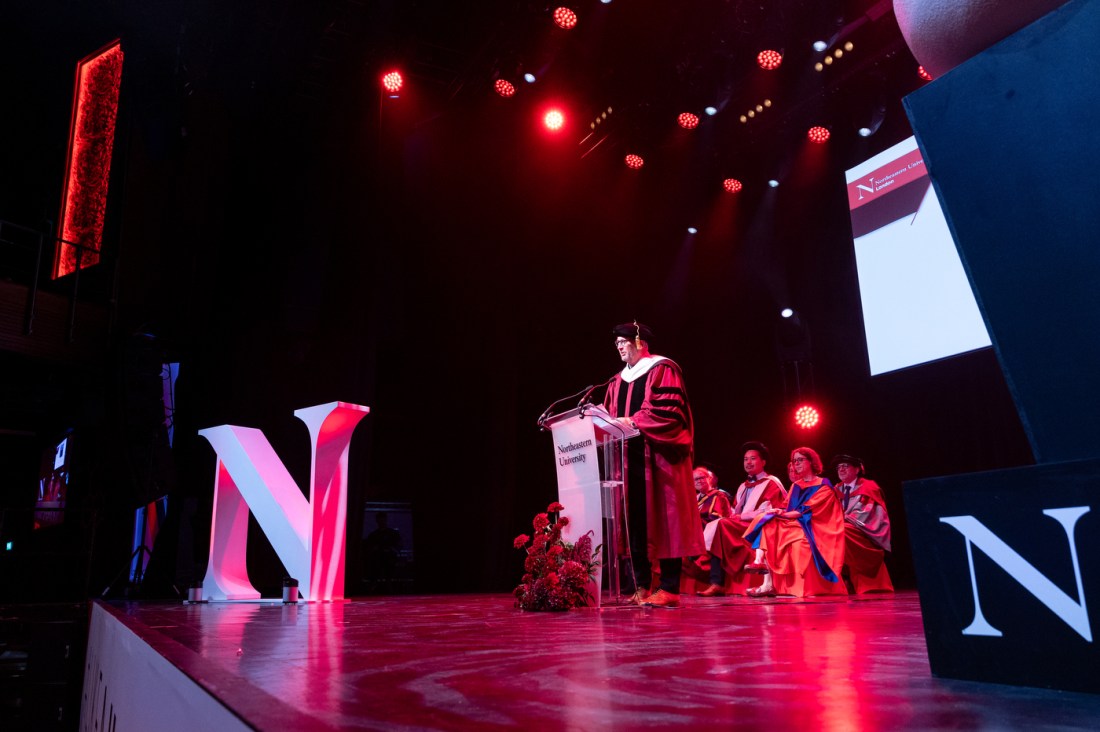 A male speaker in academic regalia addressing the crowd.