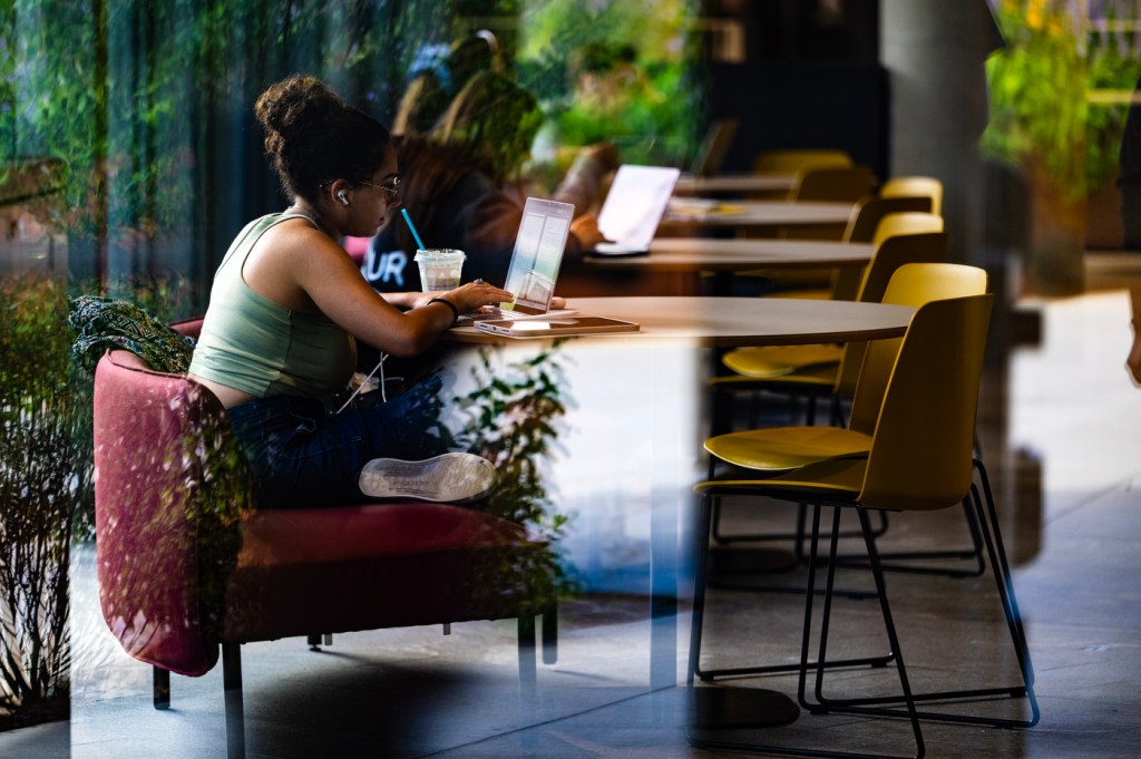 A student sits on an purple chair at a wooden table, working on a laptop computer.