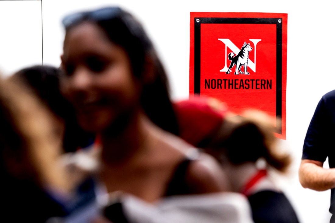A red sign with a Northeastern logo on it on a wall. In the foreground students are blurred and mingling.
