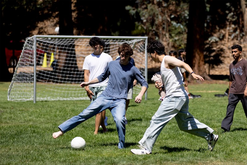 Students compete for a soccer ball on a sunny grass field at Northeastern's Oakland campus, with a goal net visible in the background.