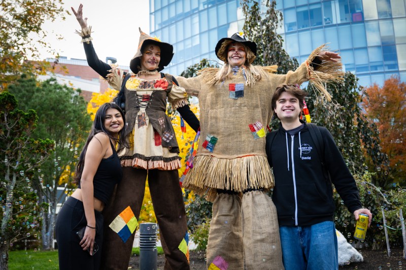 Festivalgoers pose with two people dressed as scarecrows against a backdrop of autumn trees during Northeastern’s Friends and Family Weekend Fall Festival.