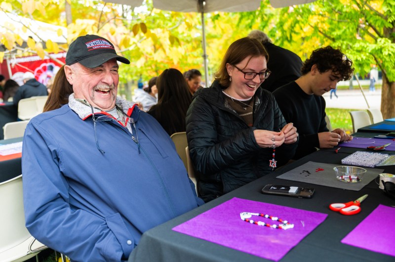 A man smiles while making friendship bracelets with family members under a tent at Northeastern’s Friends and Family Weekend Fall Festival.