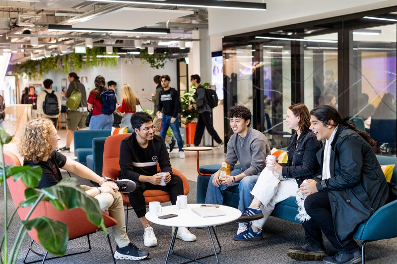 A group of students sitting in colorful, modern chairs around a low white table. They are talking and laughing amongst themselves. 