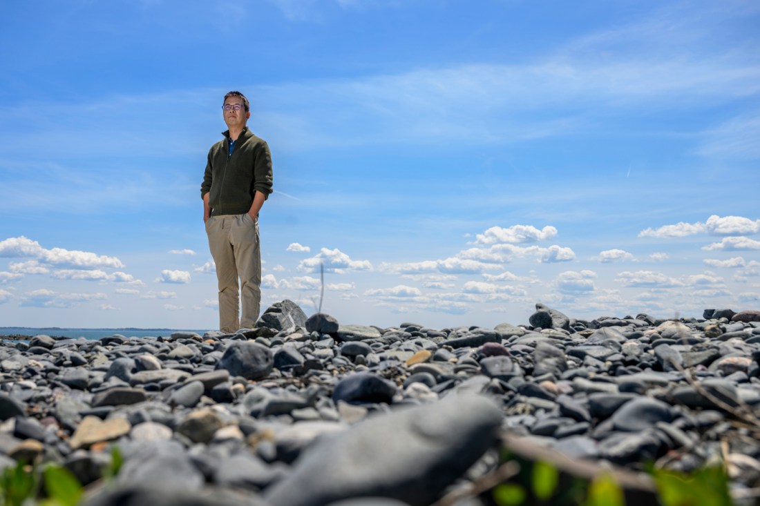 Chengfei He, an Asian man, standing on top of coastal rock formations by the sea under a blue sky.