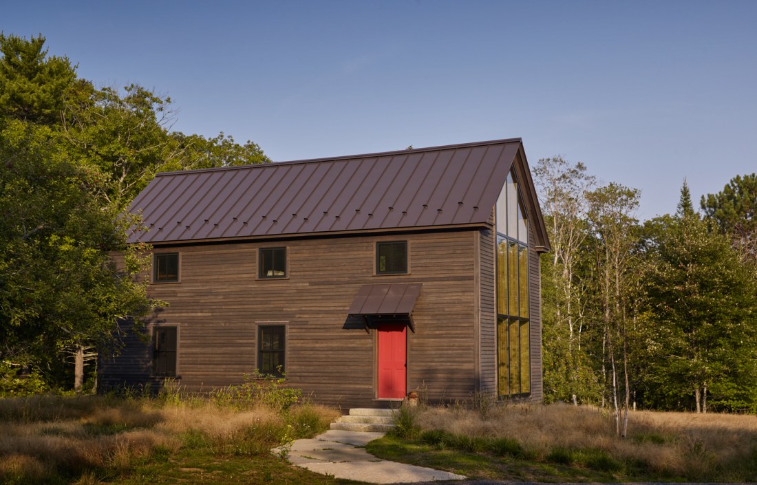 Modern barn-style house with wood siding and a dark metal roof, featuring a bright red front door and metal awning. There is a path and steps leading up to the red door.