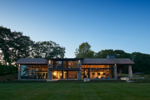 A contemporary single-story home at dusk with a stone and wood exterior and warmly lit interior spaces seen through floor-to-ceiling windows.