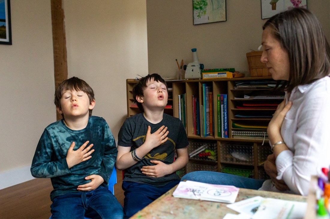 Two children and an adult practice breathing exercises together in a home setting, with their hands placed on their chests and stomachs. 